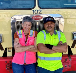 MSS coordinator Renee and driver Rafael stand in front of a garbage truck in their safety vests smiling