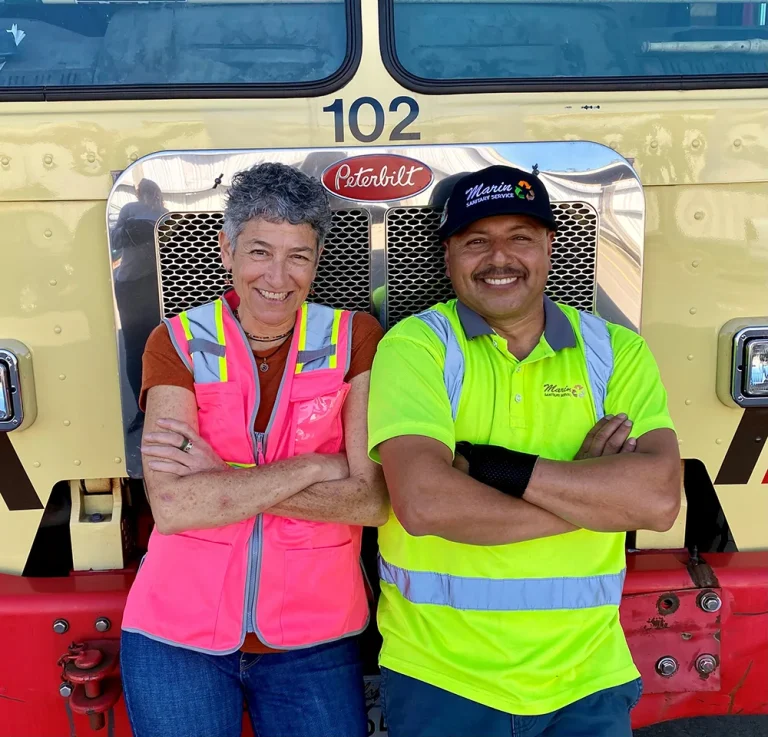 MSS Driver Rafael and Coordinator Renee stand in front of a garbage truck smiling with their safety vests on.