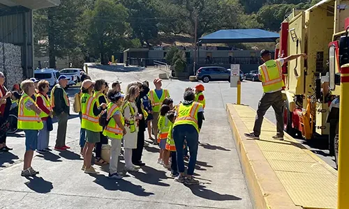A tour group in yellow jackets listens to a tour guide in front of an MSS garbage truck.